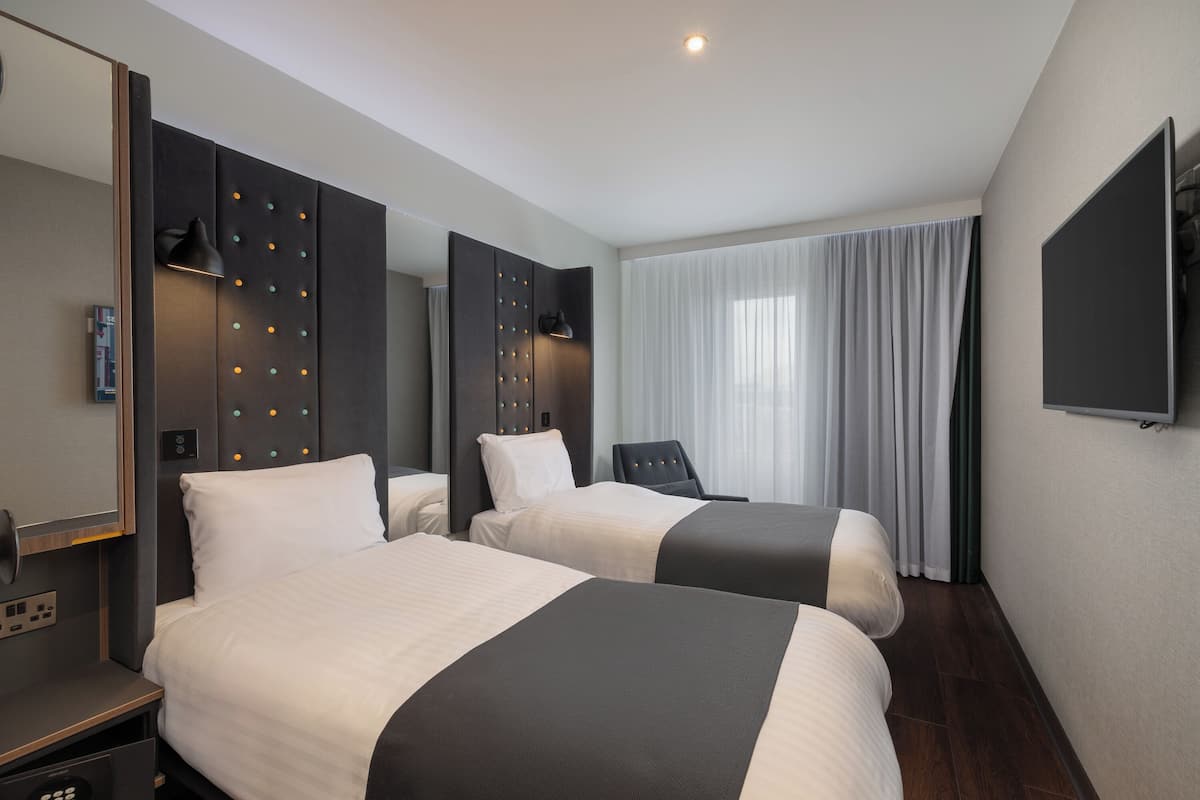 A hotel room with two beds, a TV, a desk, and a large window with curtains.