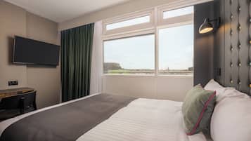 A hotel room with a bed, a desk, a television, a window with a view of a cityscape, and a wall-mounted light fixture.