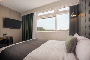 A hotel room with a bed, a desk, a television, a window with a view of a cityscape, and a wall-mounted light fixture.