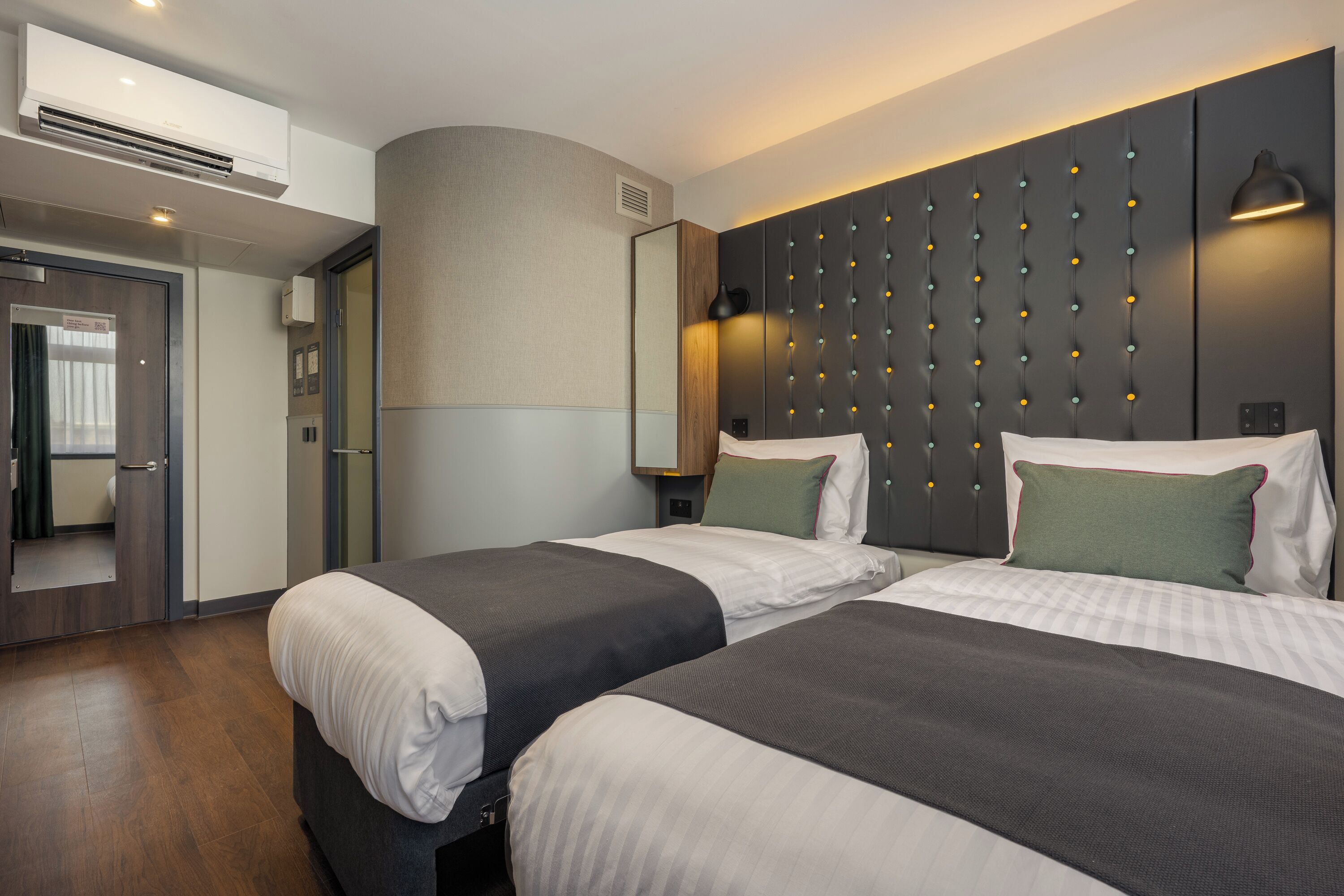 Habitación de hotel con dos camas, un espejo grande, una lámpara de pared y aire acondicionado integrado.