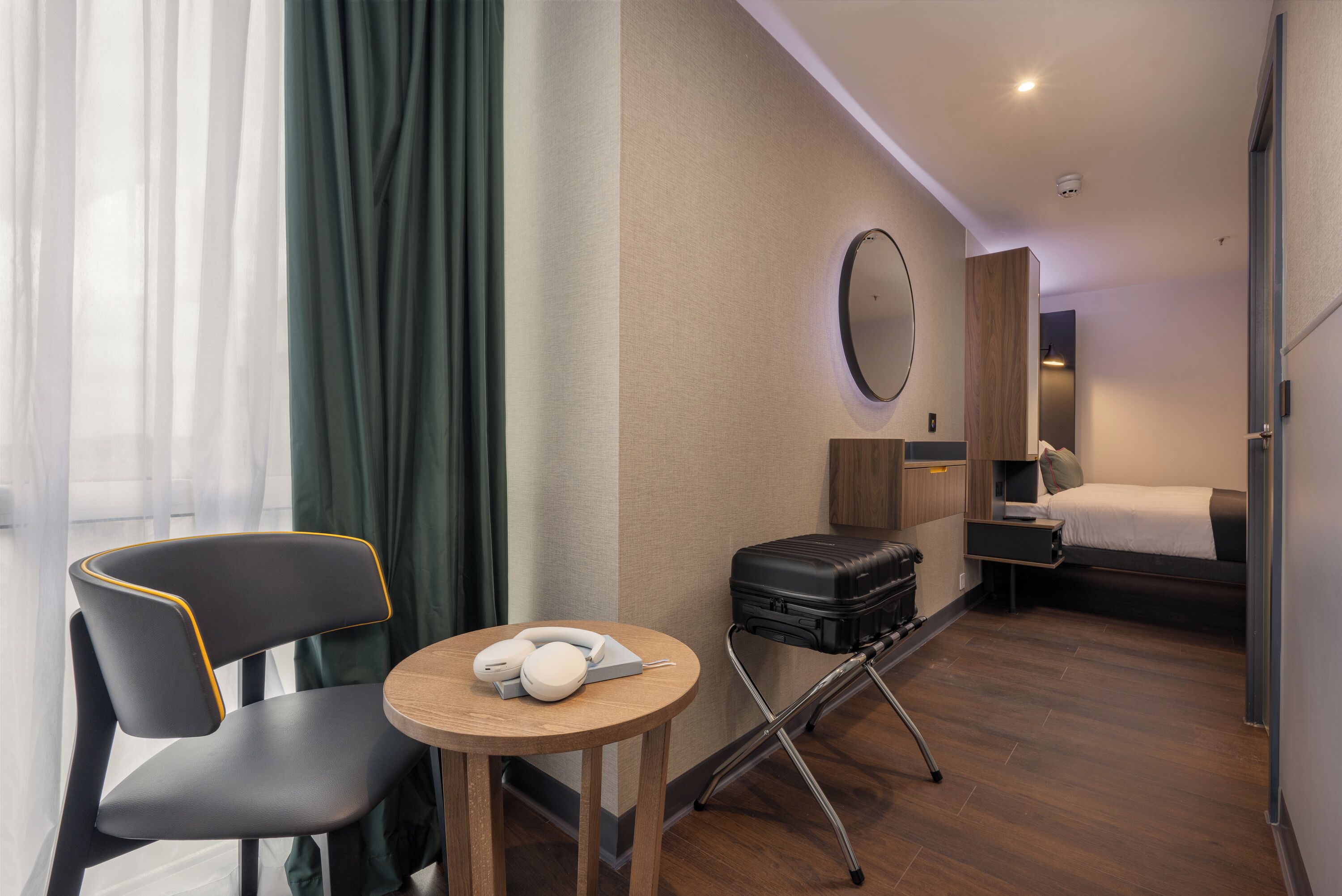 Habitación de hotel con una cama, un escritorio con silla, una pequeña mesa con otra silla, una maleta y un espejo en la pared.