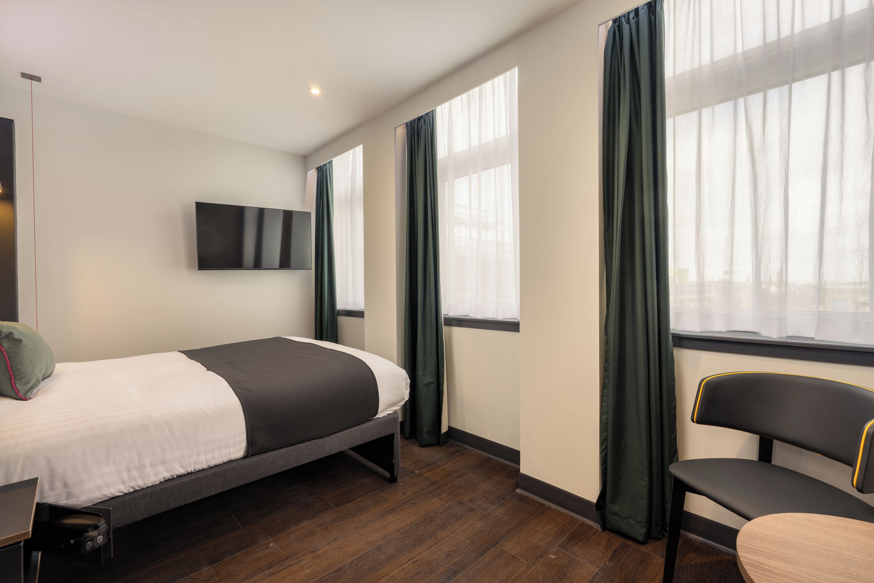 A hotel room with a bed, a chair, a TV, and windows with curtains.