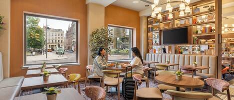 A modern lounge area with seating, a large window offering a city view, and a bookshelf with various items.
