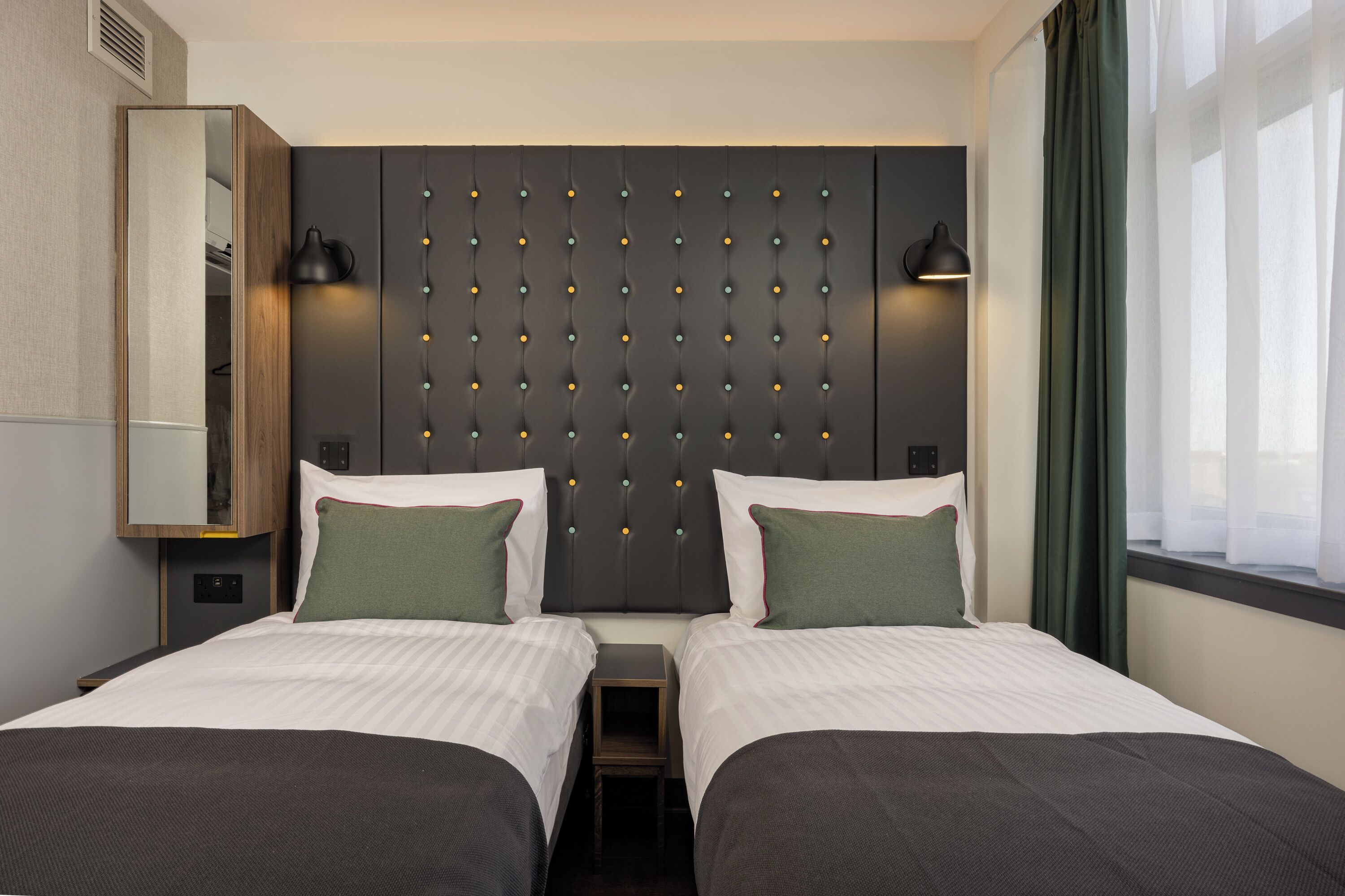 Hotelkamer met twee bedden, een grote spiegel, een hoofdeinde met een verlichtingspaneel en een raam met gordijnen.