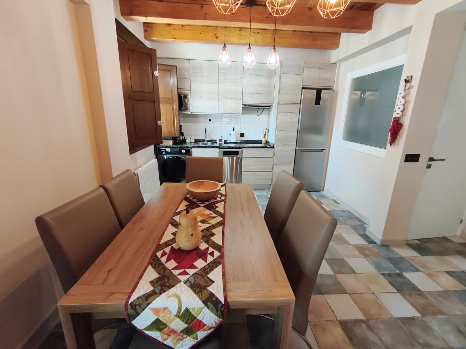 A dining area with a wooden table and chairs, a kitchen in the background, and a patterned rug on the floor.