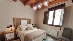 Een slaapkamer met een houten plafond, een bed met bloemenbedrukking, een nachtkastje, een stoel en een raam met zonwering.