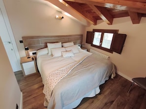 Une chambre avec un plafond en bois, un lit recouvert de plusieurs coussins, une table de chevet et une fenêtre avec des volets.