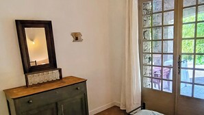 A bedroom with a bed, a dresser with a mirror, a window with a view of outdoor furniture, and a door with a curtain. - Garden apartment in the countryside near the sea pets welcome (Les Adrets-de-l'Estérel)