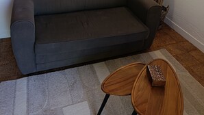 A dark gray sofa, a round wooden side table, a small rectangular wooden box, a wall-mounted decorative light, and a tall plant with long leaves. - Garden apartment in the countryside near the sea pets welcome (Les Adrets-de-l'Estérel)