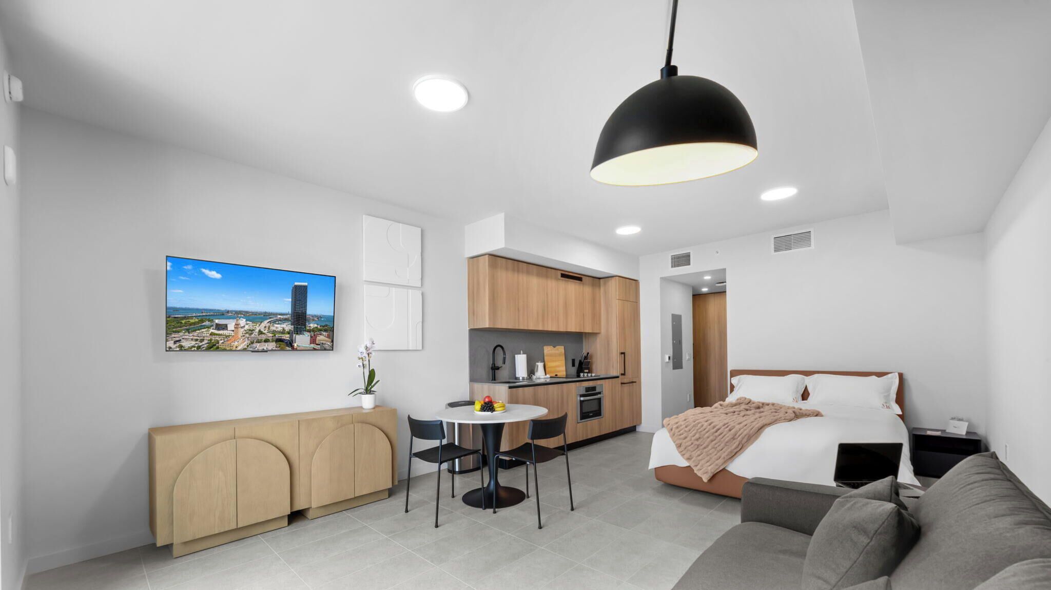 A modern hotel room with a bed, a sofa, a dining table with chairs, a kitchen area, and a cityscape view on the wall.
