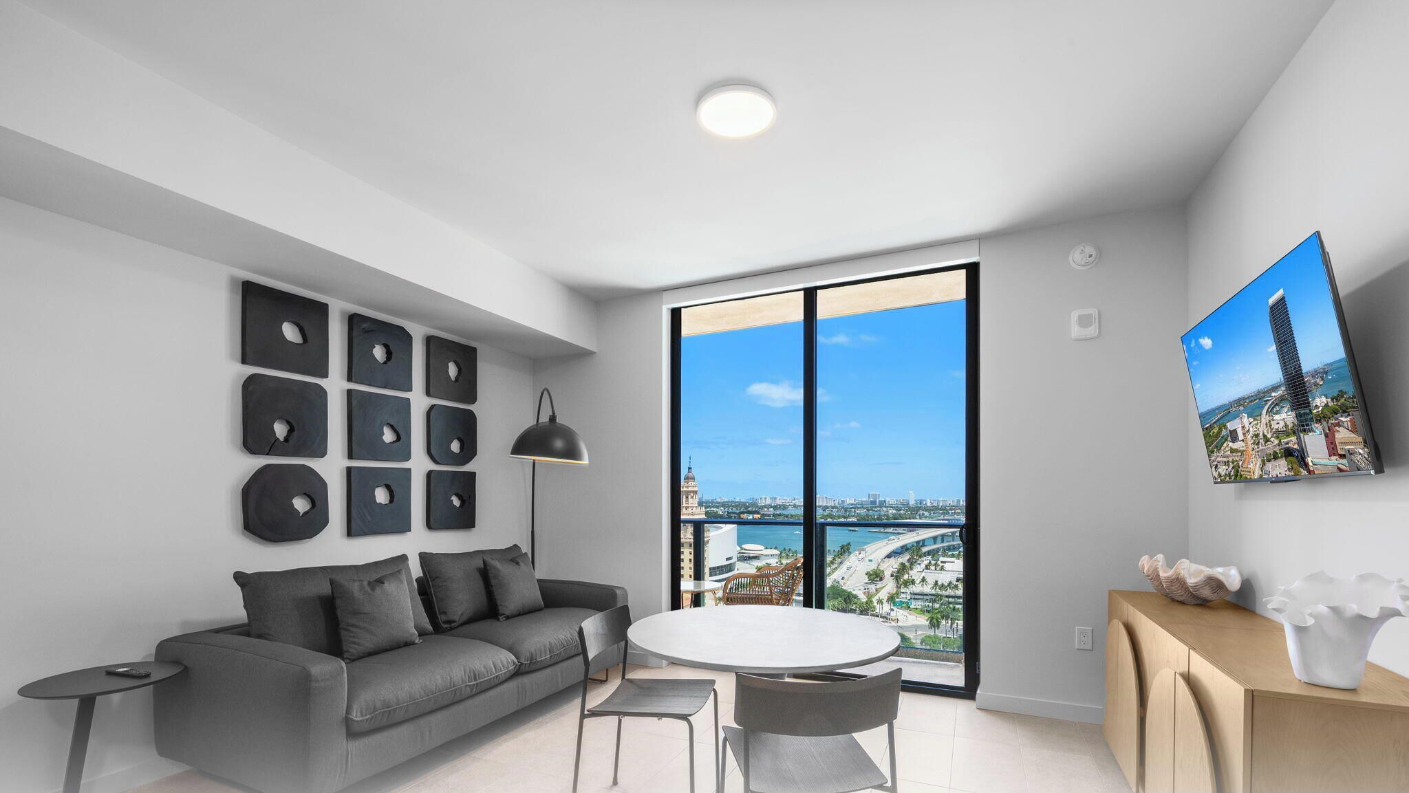 Ein modernes Wohnzimmer mit einem grauen Sofa, einem runden Tisch, Stühlen, einem großen Fenster mit Blick auf die Stadt und einem an der Wand montierten Fernseher.