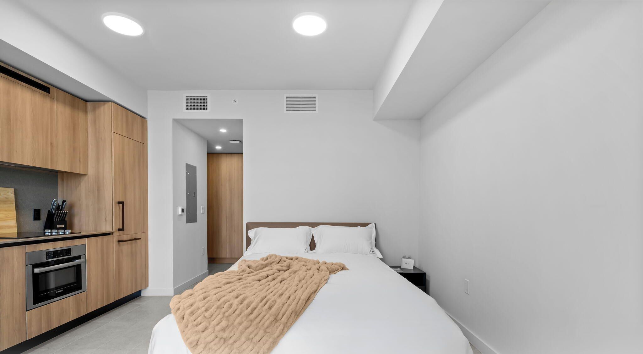 A modern hotel room with a bed, a bedside table, a kitchen area with wooden cabinets, and a built-in oven.