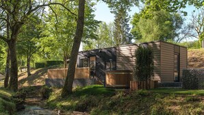 A modern log cabin with a wooden deck, a hot tub, and a stream in the foreground.