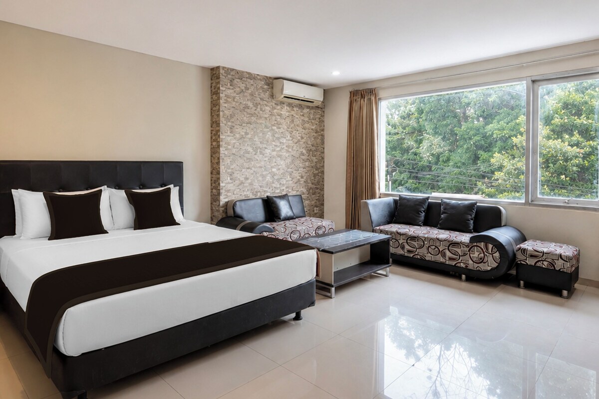 A hotel room with a bed, a sofa, a small table, and a window with a view of trees.