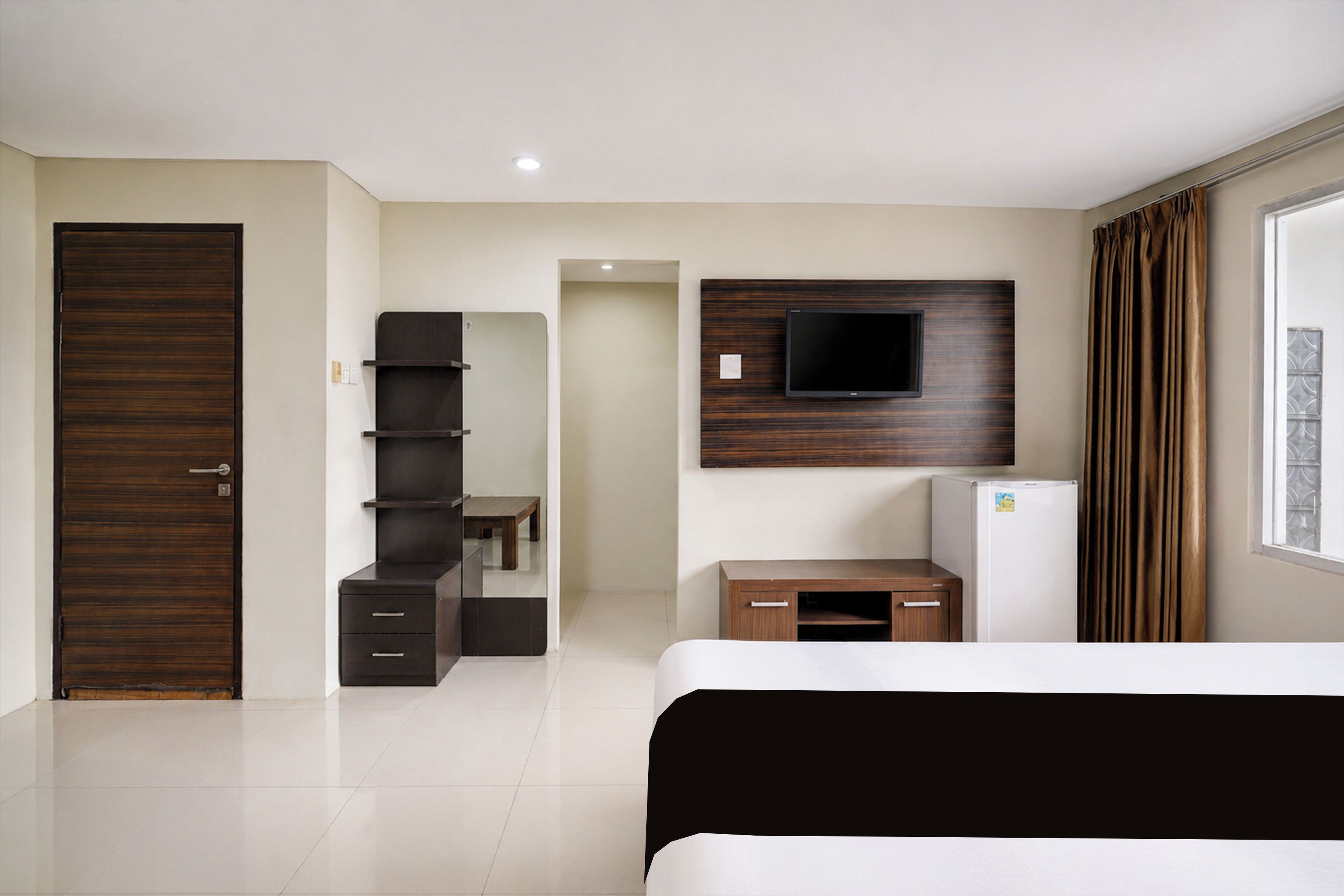 A modern hotel room with a bed, a TV mounted on the wall, a small kitchenette, and a wardrobe.