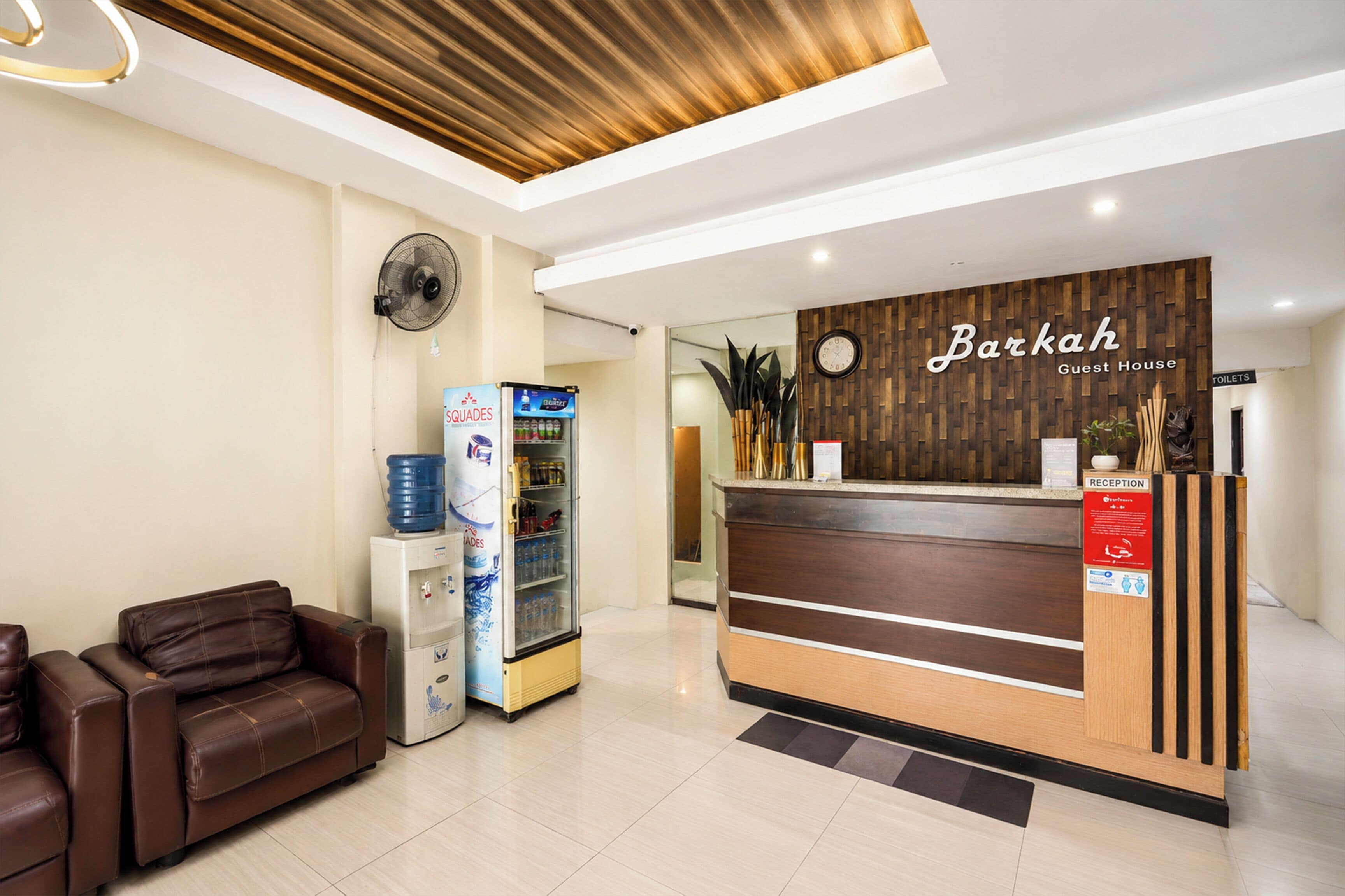 A reception desk with a wooden panel design, a water dispenser, a fan, and a reception area with a brown leather sofa.