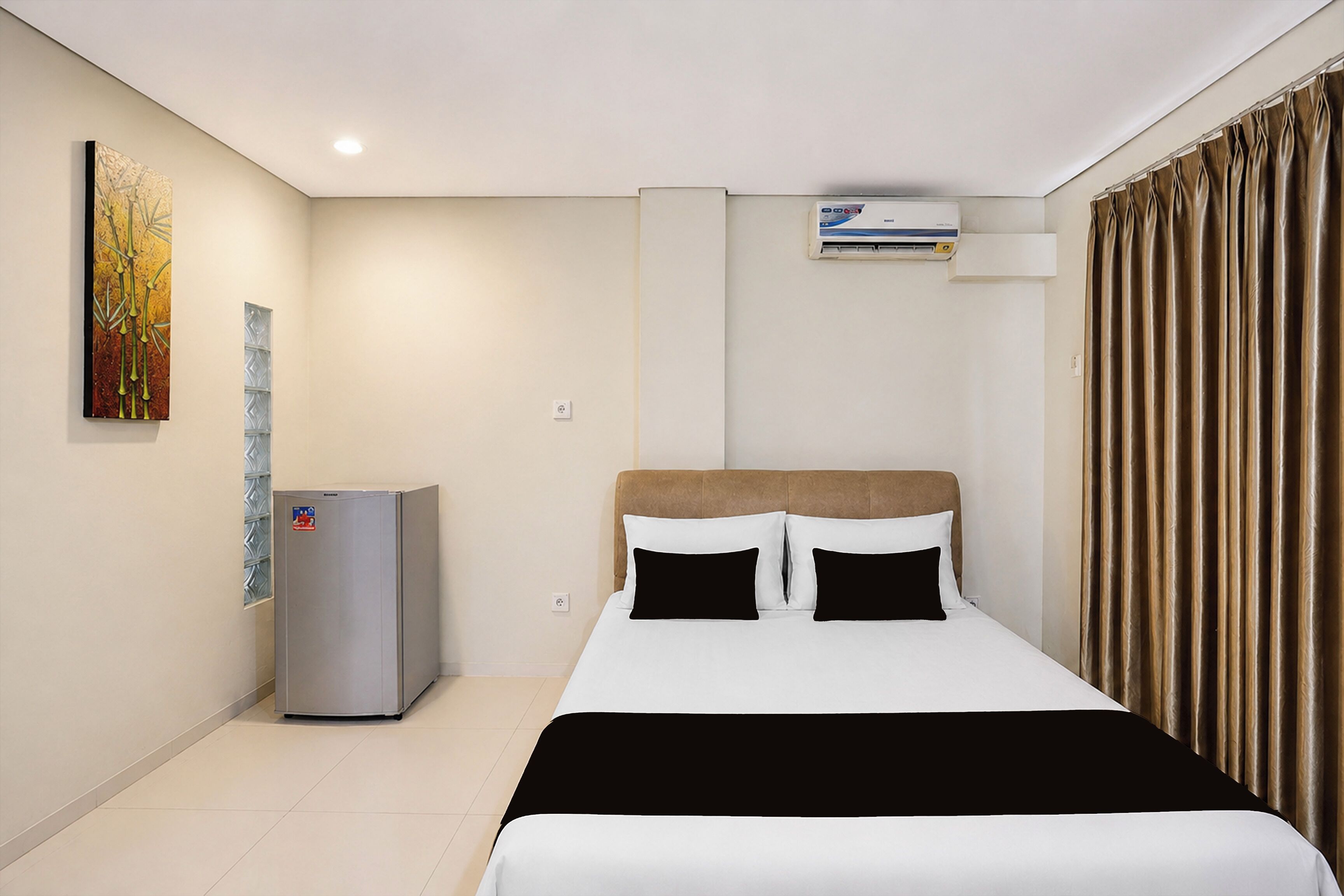 A hotel room with a bed, a wall-mounted air conditioner, a refrigerator, and a window with curtains.