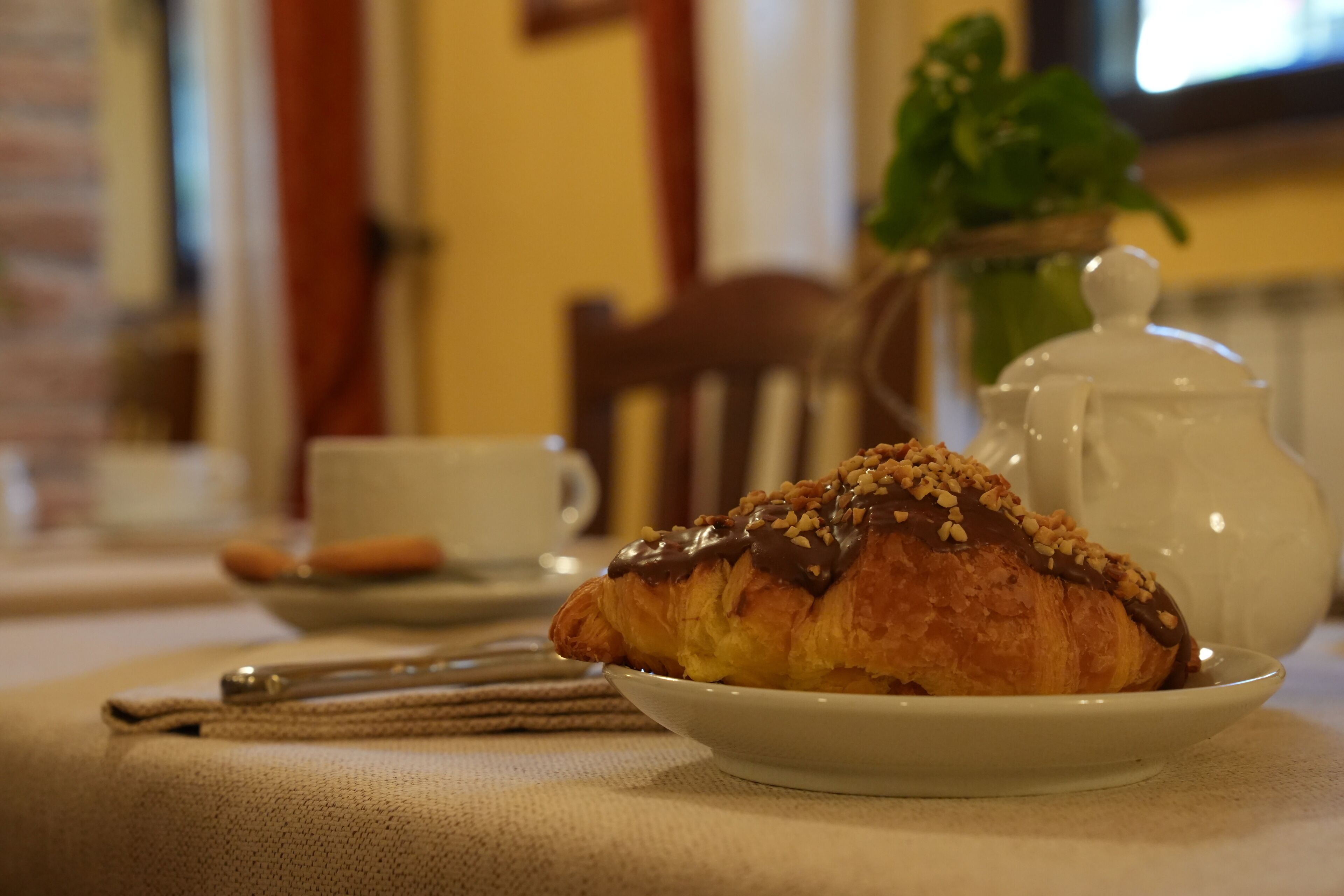 A croissant on a plate with a teapot and a cup in the background.
