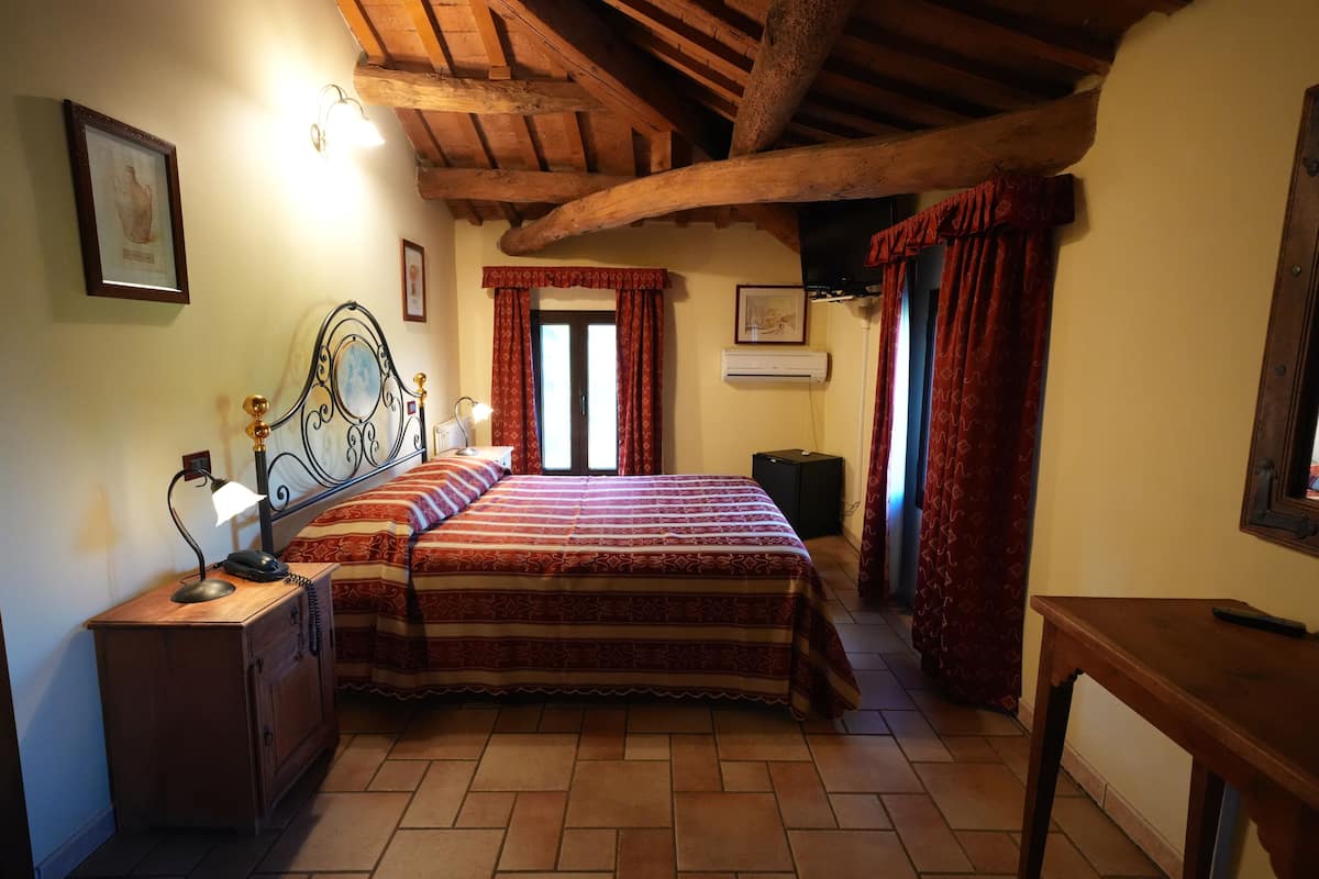 Un dormitorio con techo de madera, cama con colcha estampada, mesita de noche con lámpara y ventana con cortinas rojas.