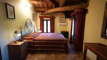 Quarto com teto de madeira, cama com colcha estampada, mesa de cabeceira com abajur e janela com cortinas vermelhas.