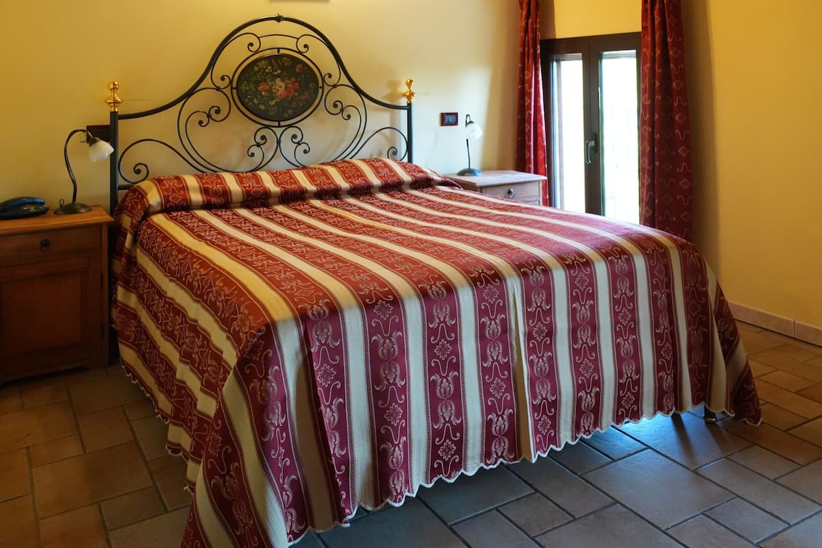 Un dormitorio con techo de madera, una cama con colcha estampada, una mesita de noche, una lámpara de pared y una ventana con cortinas.