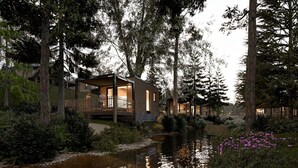 A modern cabin-style building surrounded by dense forest, with a small pond and flowering plants in the foreground.