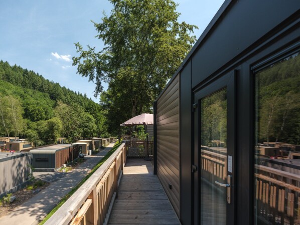 Une terrasse en bois mène à un bâtiment doté de grandes fenêtres, qui est lui-même entouré d’une zone boisée.