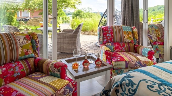 Une pièce baignée de lumière, avec deux fauteuils colorés, une petite table sur laquelle sont posés des verres et des bouteilles de vin, et une vue sur un jardin et des collines à l’extérieur.