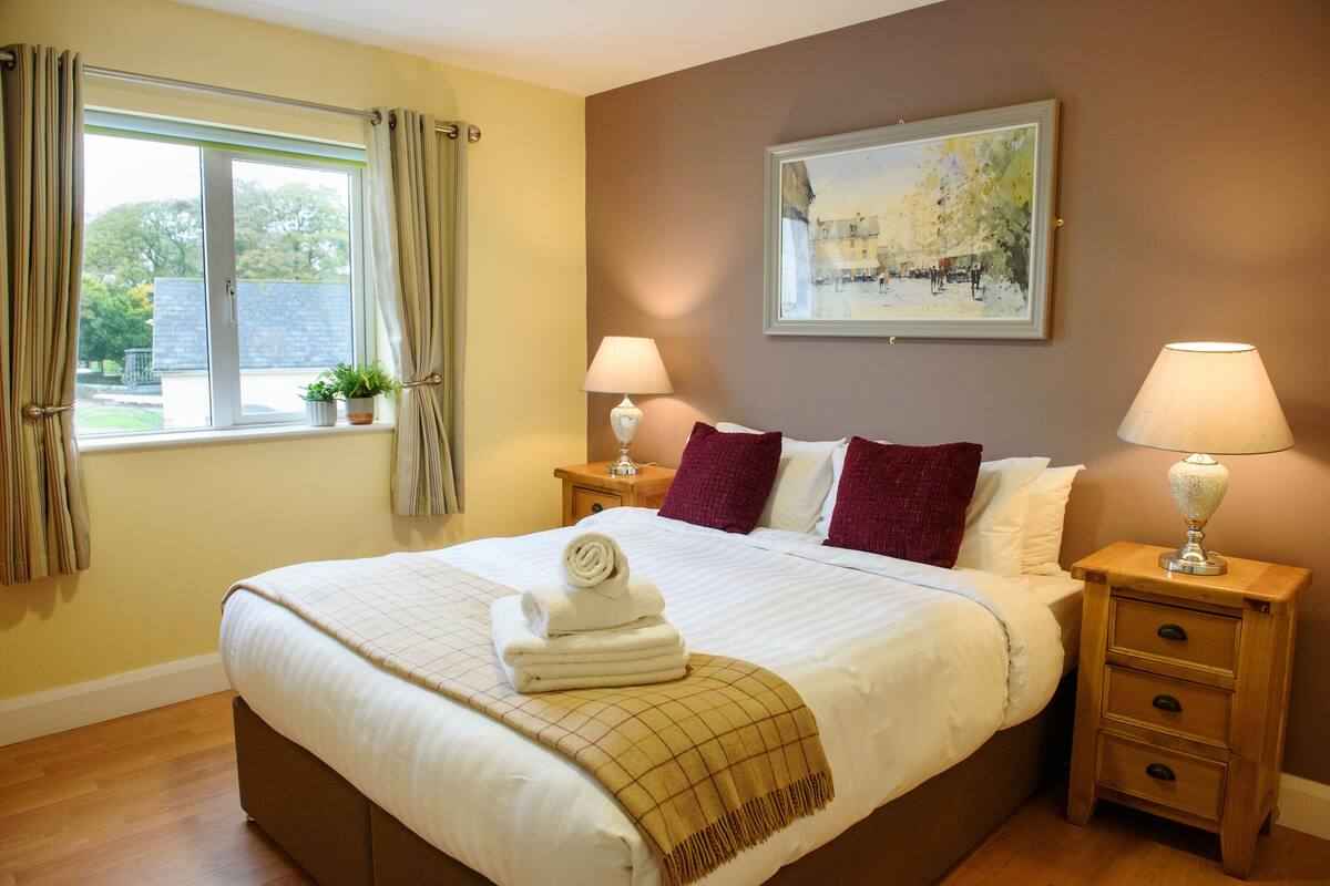 A neatly made bed with a rolled towel, two bedside lamps, a window with curtains, and a painting on the wall.