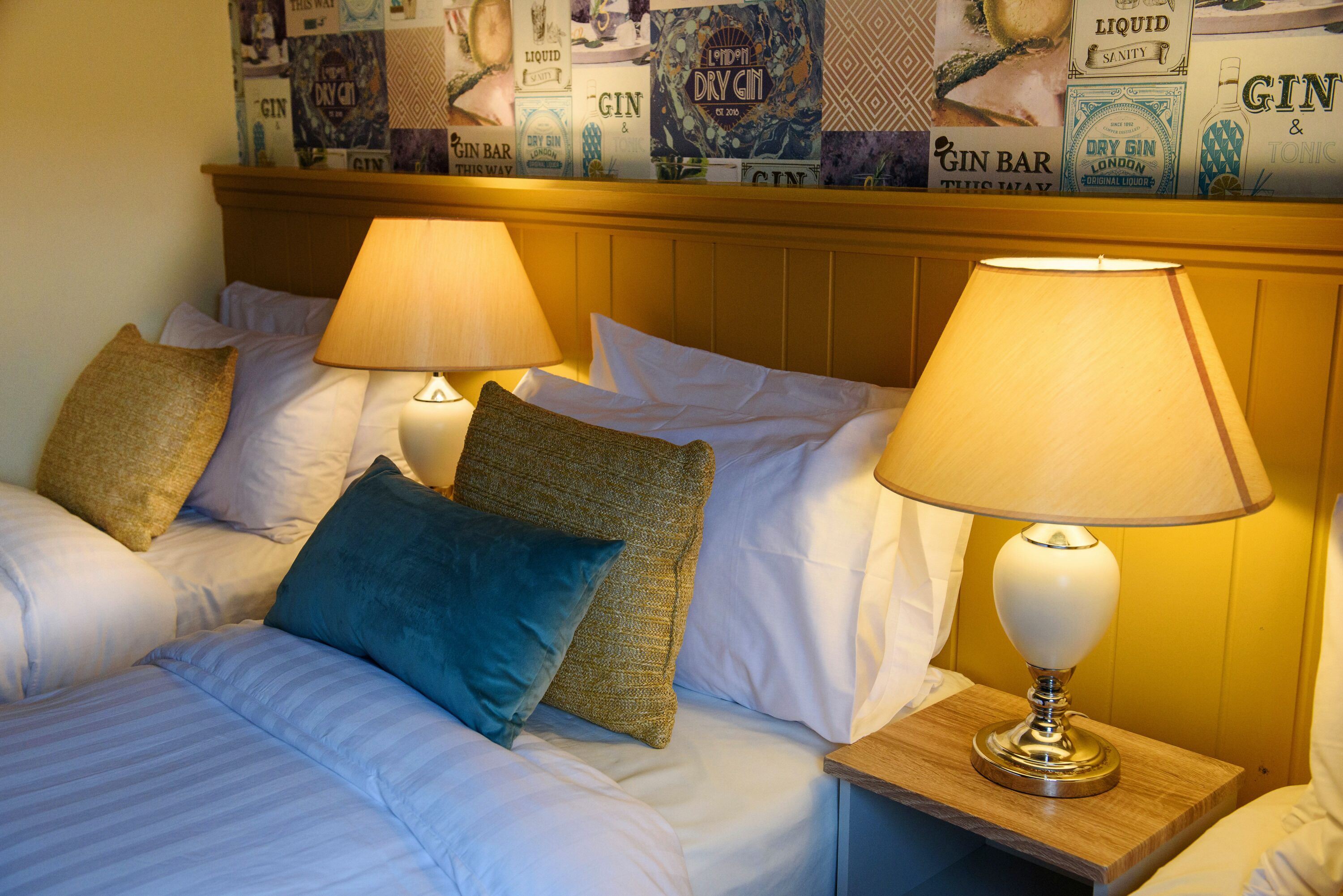 A hotel room with two beds, each with a lamp, pillows, and a wall with gin advertisements.