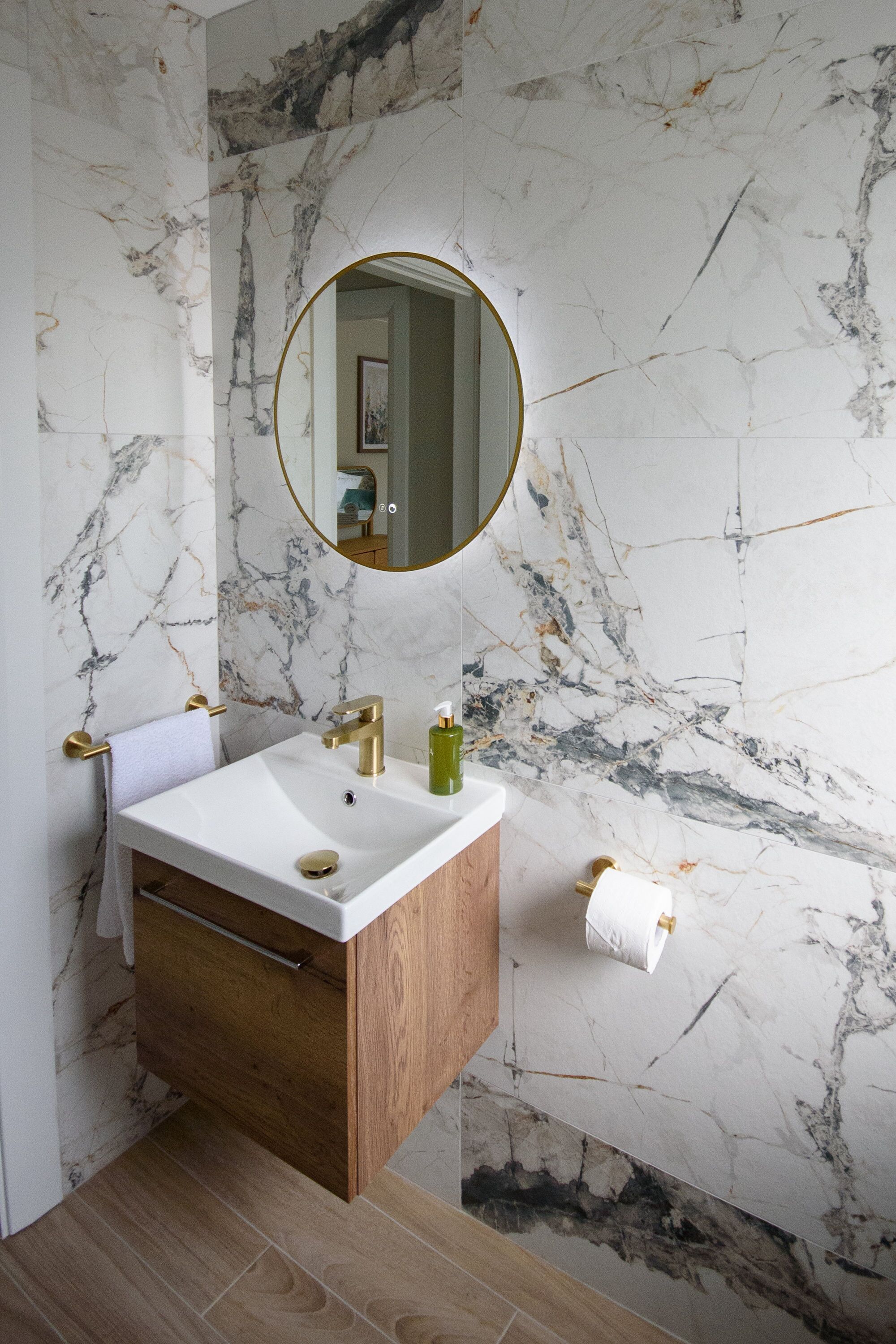 A bathroom with a marble wall, a wooden vanity, a gold faucet, and a round mirror.