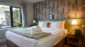 A neatly made bed with white linens and a rolled towel, a bedside lamp, and a small plant on a wooden nightstand.