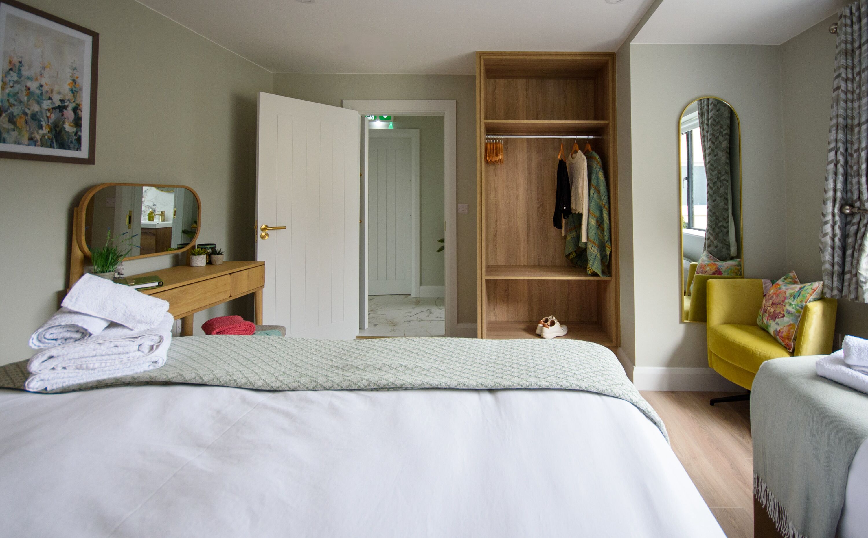 A bedroom with a bed, a wooden wardrobe, a mirror, a yellow armchair, and a painting on the wall.
