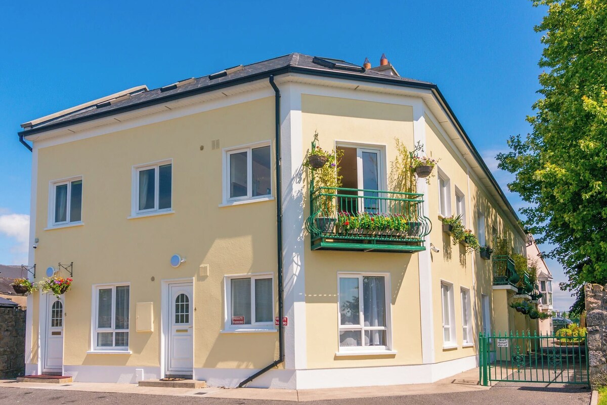A two-story building with a flat roof, a balcony with hanging flower baskets, and a gated entrance.