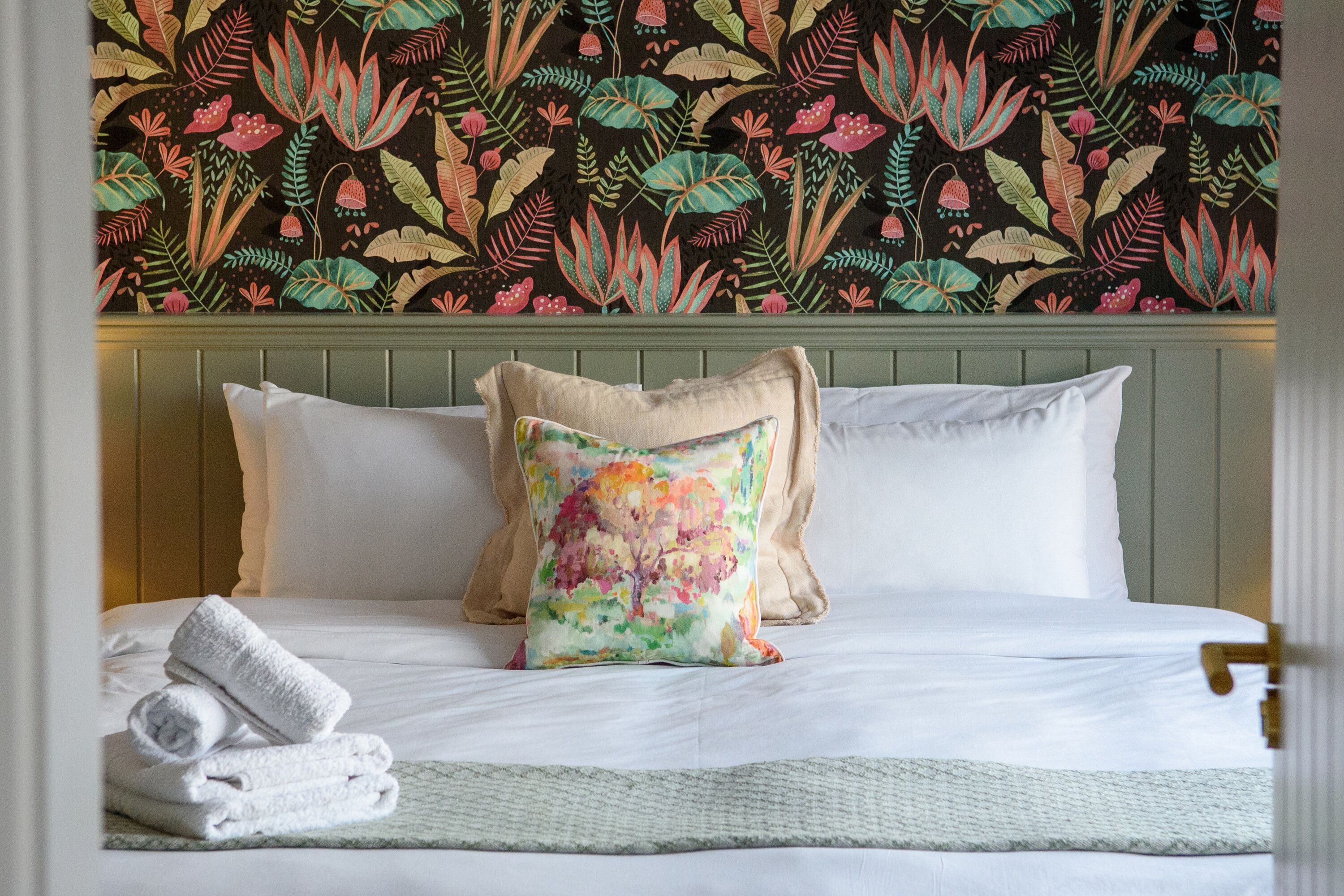 A neatly made bed with white linens and a colorful floral pillow, set against a backdrop of a patterned wallpaper.