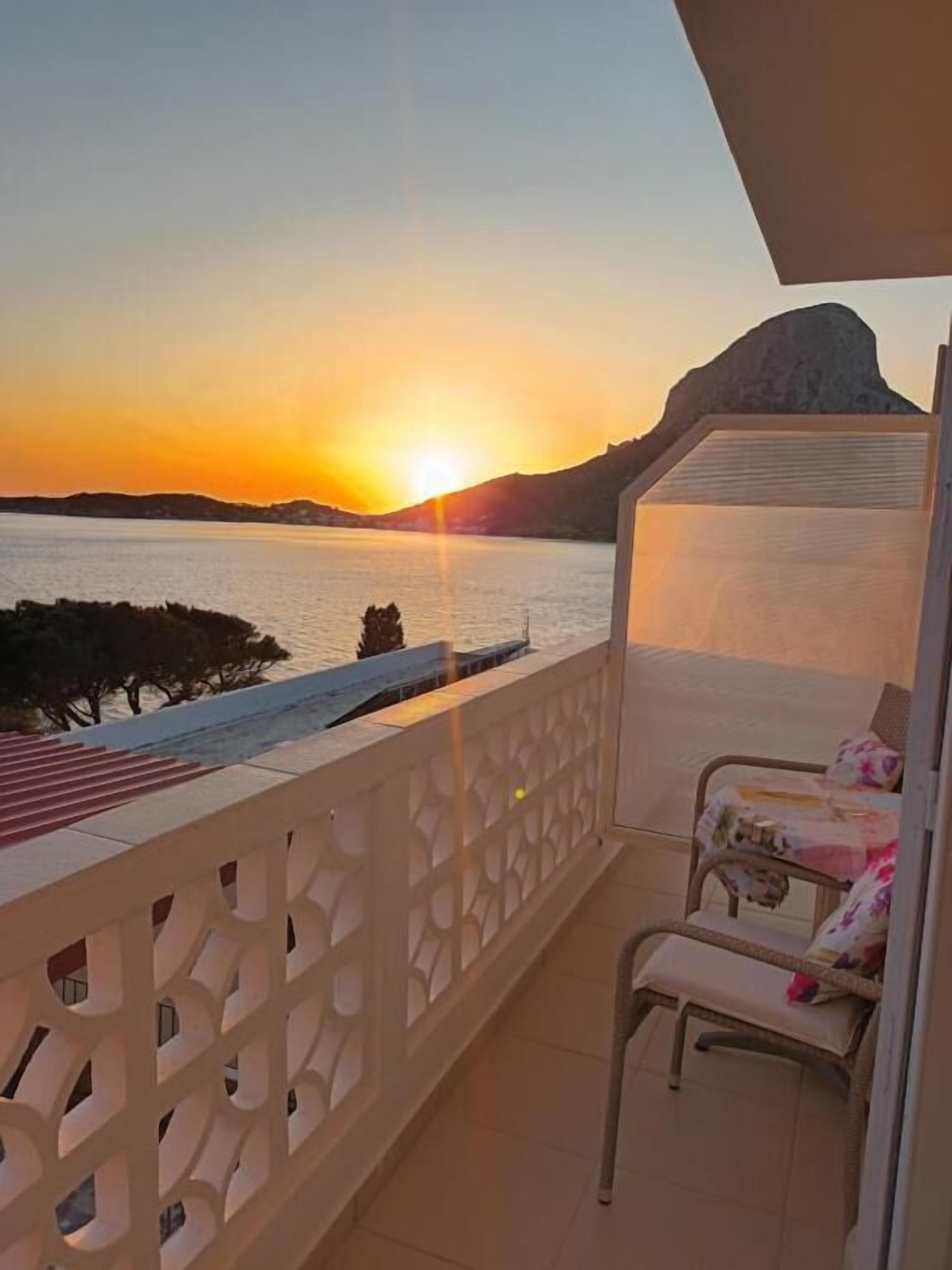 A balcony with a view of the sunset over water, a chair, and a bed with a patterned bedspread.