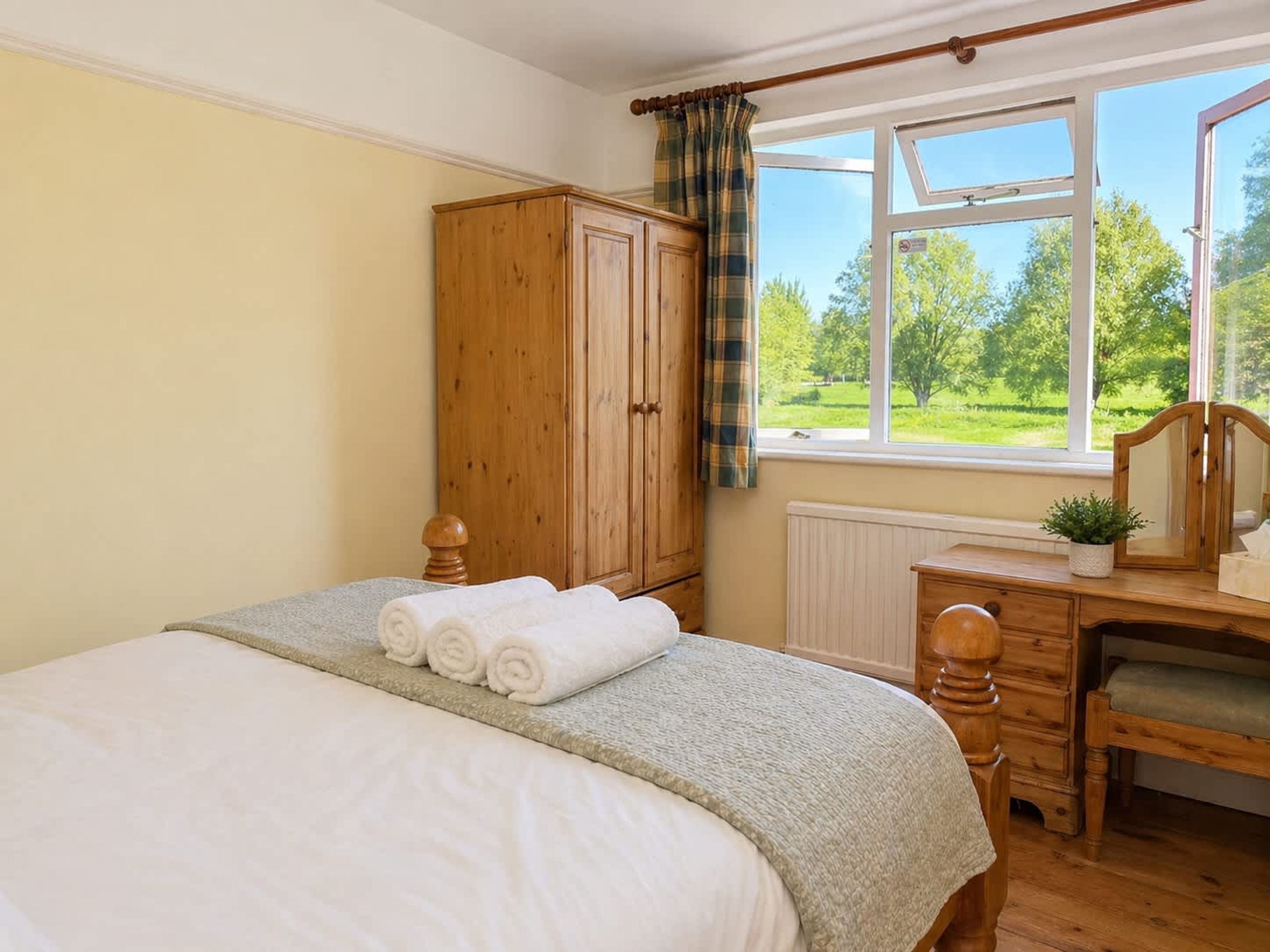 Un dormitorio con cama, armario de madera, escritorio con espejo y ventana con vista a vegetación.