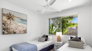 Ein Schlafzimmer mit zwei Betten, einem Deckenventilator, einem Fenster mit Blick auf Grünflächen und einem Bild von einer Strandlandschaft.