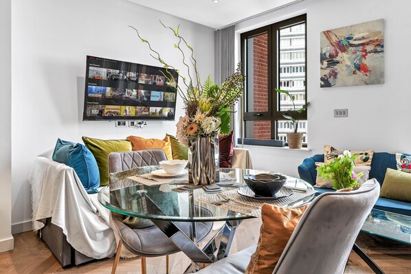 Ein modernes Wohnzimmer mit Glastisch, bequemen Sitzmöglichkeiten, einem Flachbildfernseher und einem großen Fenster mit Blick auf Gebäude.