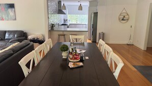 Un salón moderno con una mesa de comedor de madera oscura, sillas blancas, un sofá de cuero negro y una cocina al fondo.