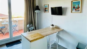 Une petite pièce avec un bureau en bois, une chaise blanche, un téléviseur fixé au mur et une porte-fenêtre coulissante donnant sur un espace extérieur aménagé avec des chaises.