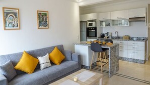 Una sala de estar moderna con un sofá gris, cojines amarillos y una mesa de centro. Se observa una cocina con gabinetes blancos y una encimera de granito al fondo.