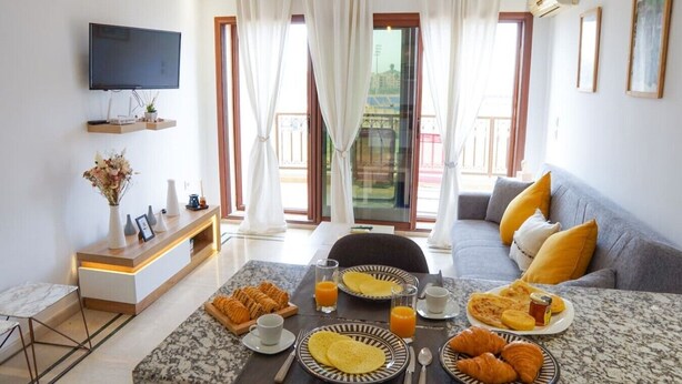 Una sala de estar moderna con un sofá, una mesa de comedor con desayuno y un televisor en la pared.