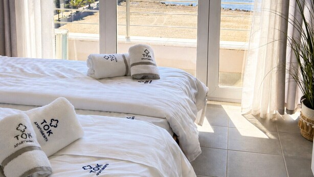 Een hotelkamer met een grote schuifglazen deur die uitkomt op uitzicht op het strand, een bed met witte lakens en twee handdoeken met de branding 'TOK APARTMENTS'.