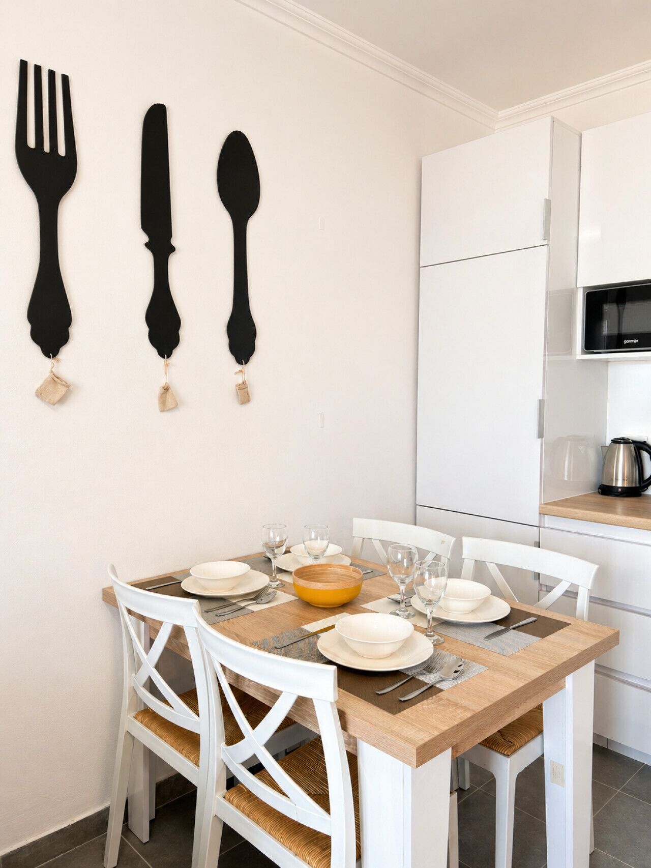 A dining area with a wooden table set for four, white chairs, and a kitchen with white cabinets and a microwave.