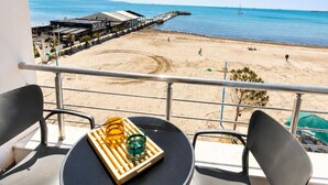 Een balkon met een tafel en stoelen met uitzicht op een strand met een pier en een zeilboot.