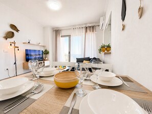 Ein Essbereich mit Tisch für vier Personen, einem Fernseher im Hintergrund und einem Fenster mit Vorhängen.