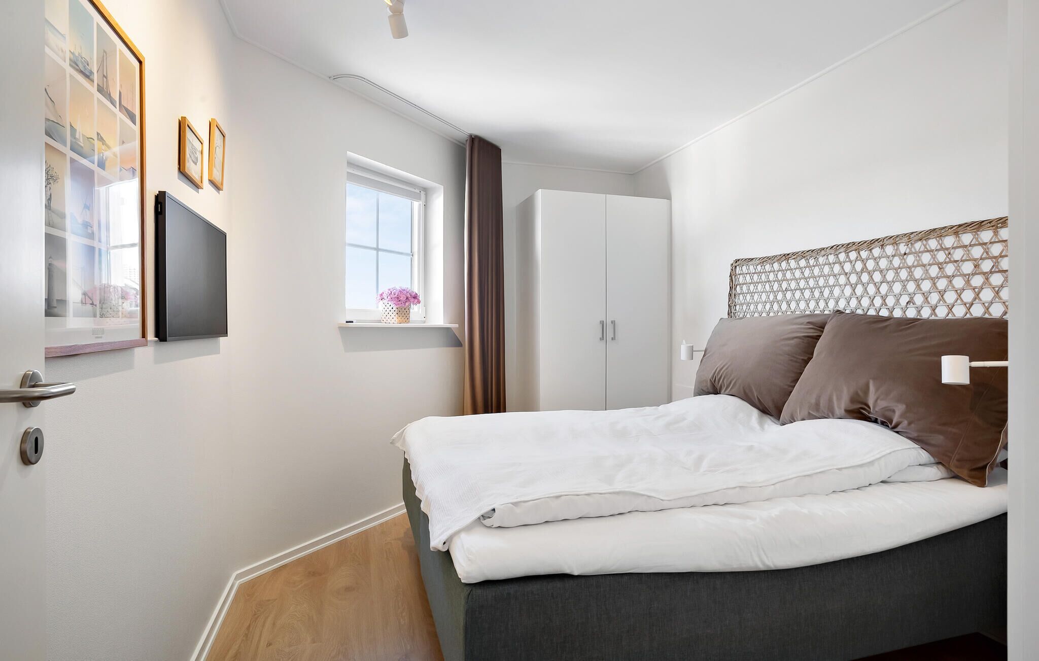 A bedroom with a bed, a wardrobe, a window with curtains, a flat-screen TV, and a wall-mounted shelf with a vase.