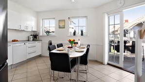 Une cuisine moderne avec des meubles blancs, une table à manger entourée de chaises noires et une vue sur l’extérieur grâce à une porte-fenêtre coulissante.