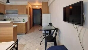 Une cuisine compacte avec des meubles en bois, une table à manger avec des chaises, un téléviseur fixé au mur et un petit coin salon agrémenté d’un coussin bleu.
